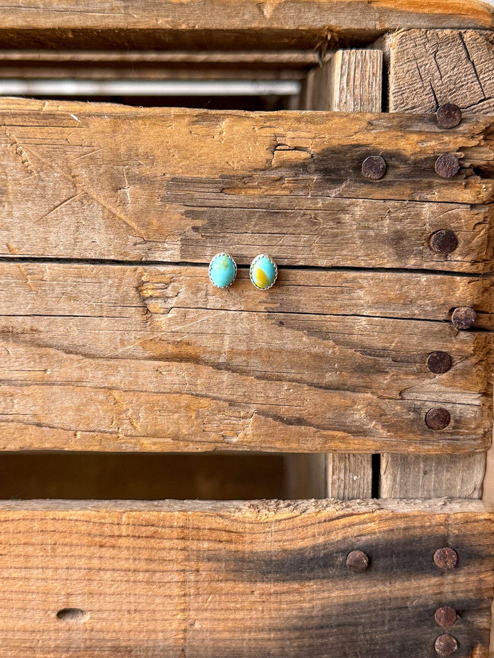 A pair of Anoka Turquoise Stud Earrings by Savannah Sevens western life{&}style, featuring small round Kingman turquoise stones set in sterling silver edges, are displayed on a rustic wooden surface with visible nails and grain.