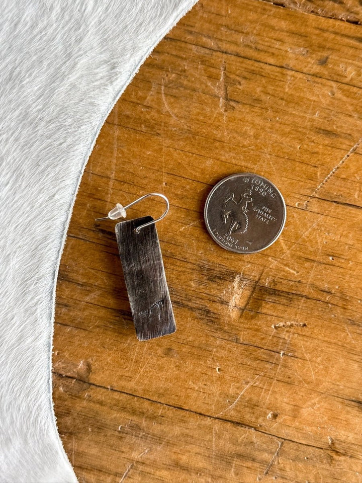 The Aquilla Earrings by Savannah Sevens western life{&}style are rectangular sterling silver earrings with a hook and clear backing, shown on wood beside a Wyoming state quarter for size. A white textured fabric appears in the upper left corner.