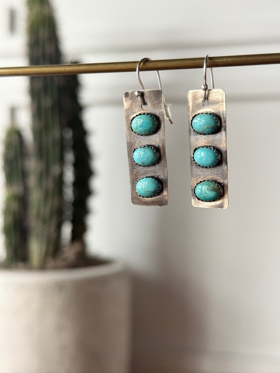 The Aquilla Earrings by Savannah Sevens western life{&}style feature a rectangular sterling silver design with three round Kingman turquoise stones on each, suspended from a rod. A blurred tall cactus in a white pot is in the background.