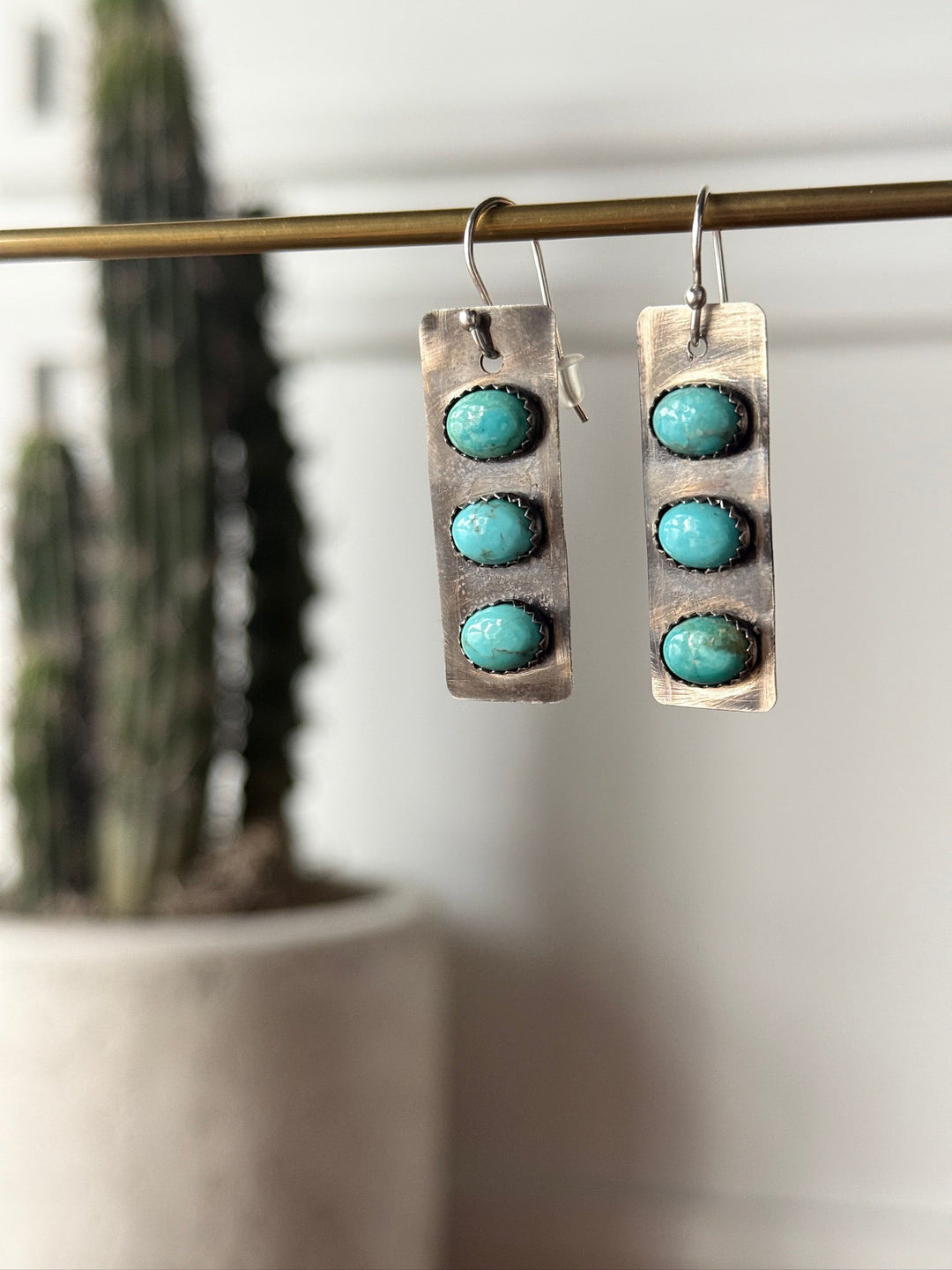 The Aquilla Earrings by Savannah Sevens western life{&}style feature a rectangular sterling silver design with three round Kingman turquoise stones on each, suspended from a rod. A blurred tall cactus in a white pot is in the background.