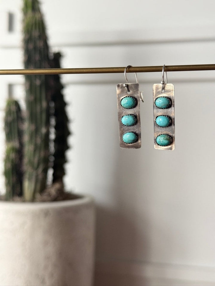 The Aquilla Earrings by Savannah Sevens western life{&}style are rectangular sterling silver earrings, each with three round Kingman turquoise stones dangling from a metal rod, shown with a blurred tall potted cactus in the background.