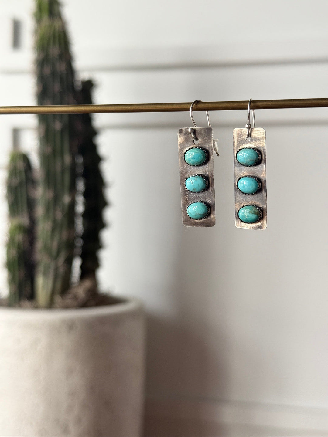 The Aquilla Earrings by Savannah Sevens western life{&}style are rectangular sterling silver earrings, each with three round Kingman turquoise stones dangling from a metal rod, shown with a blurred tall potted cactus in the background.