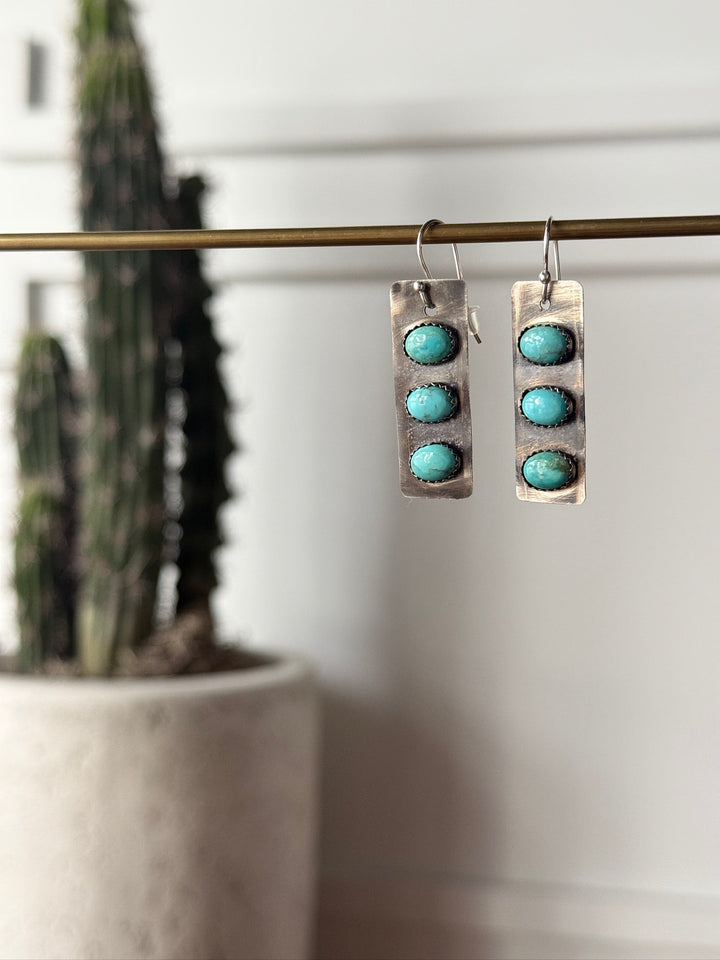 The Aquilla Earrings by Savannah Sevens western life{&}style are rectangular sterling silver earrings, each with three round Kingman turquoise stones dangling from a metal rod, shown with a blurred tall potted cactus in the background.