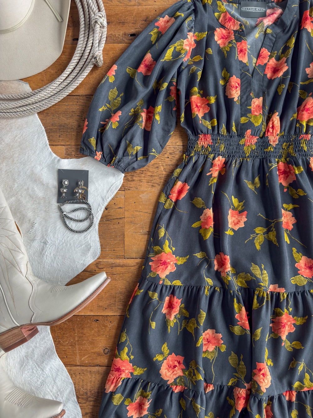 Floral print midi dress with red and yellow flowers, featuring a shirred elastic waistline, paired with silver jewelry, white cowboy boots, a white hat, and a coiled rope arranged on a wooden floor.