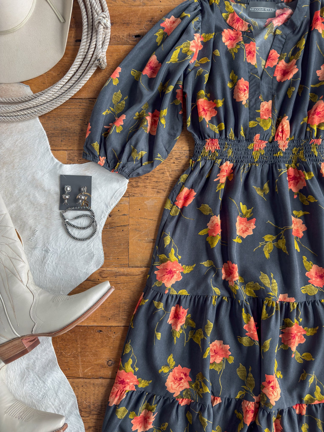 Floral print midi dress with red and yellow flowers, featuring a shirred elastic waistline, paired with silver jewelry, white cowboy boots, a white hat, and a coiled rope arranged on a wooden floor.