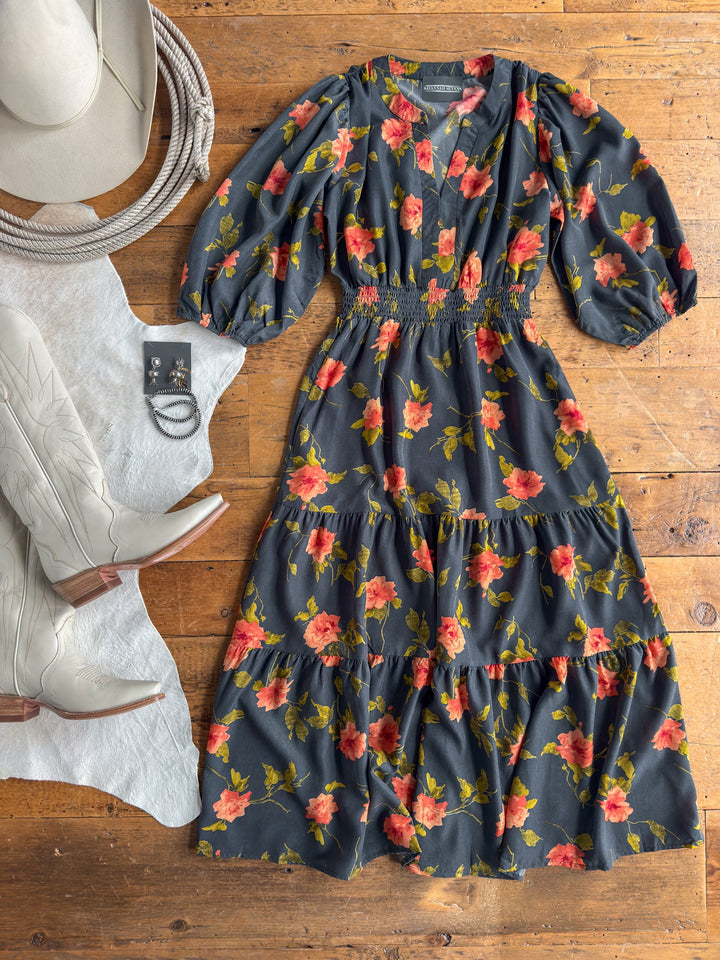 The Arcata Slate Floral Half Sleeve Dress by Savannah Sevens western life{&}style, featuring a dark floral print with red and orange blooms and a shirred elastic waistline, is laid out on a wooden floor alongside a white cowboy hat, silver jewelry, and off-white cowboy boots.