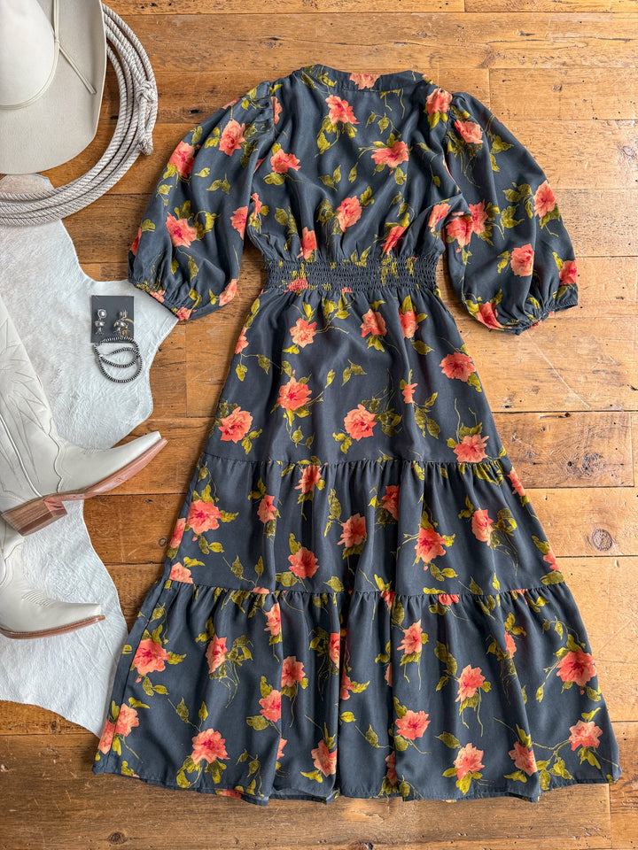 The Arcata Slate Floral Half Sleeve Dress by Savannah Sevens western life{&}style is displayed on a wooden floor, surrounded by a white hat, white boots, earrings, and bracelets. This dark midi dress features a vibrant  floral print and is designed with half sleeves and a shirred elastic waistline for added comfort and style.