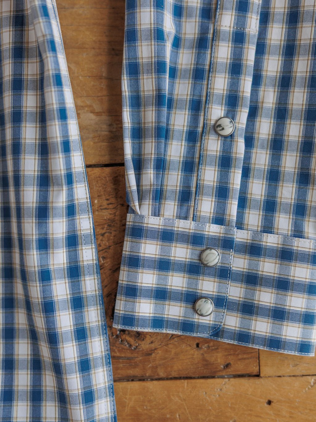Close-up of the Ariat Howell Men's Plaid Pearl Snap by Savannah Sevens western life{&}style, featuring blue, white, and beige plaid cuffs with two white buttons, laid on wood. Classic style meets modern comfort in this men's shirt.