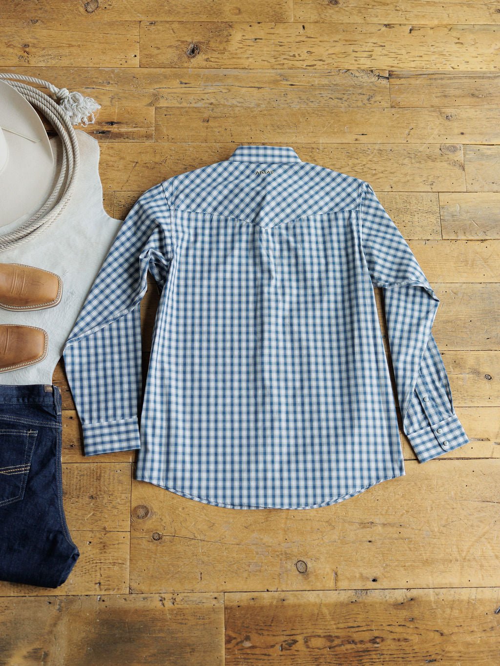 The Ariat Howell Men's Plaid Pearl Snap shirt by Savannah Sevens western life{&}style is laid flat on a wooden floor, surrounded by blue jeans, tan shoes, a white hat, and a white cloth.