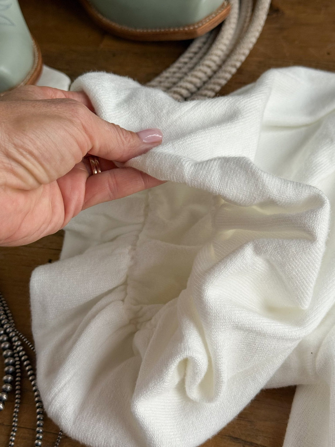 A hand holds the Arietta White Puff Sleeve Top by Savannah Sevens western life{&}style, showcasing its white textured fabric and gathered elastic stitching. In the background, a green boot, coiled rope, and beaded necklace rest on a wooden surface, highlighting the intricate details of this stylish top.