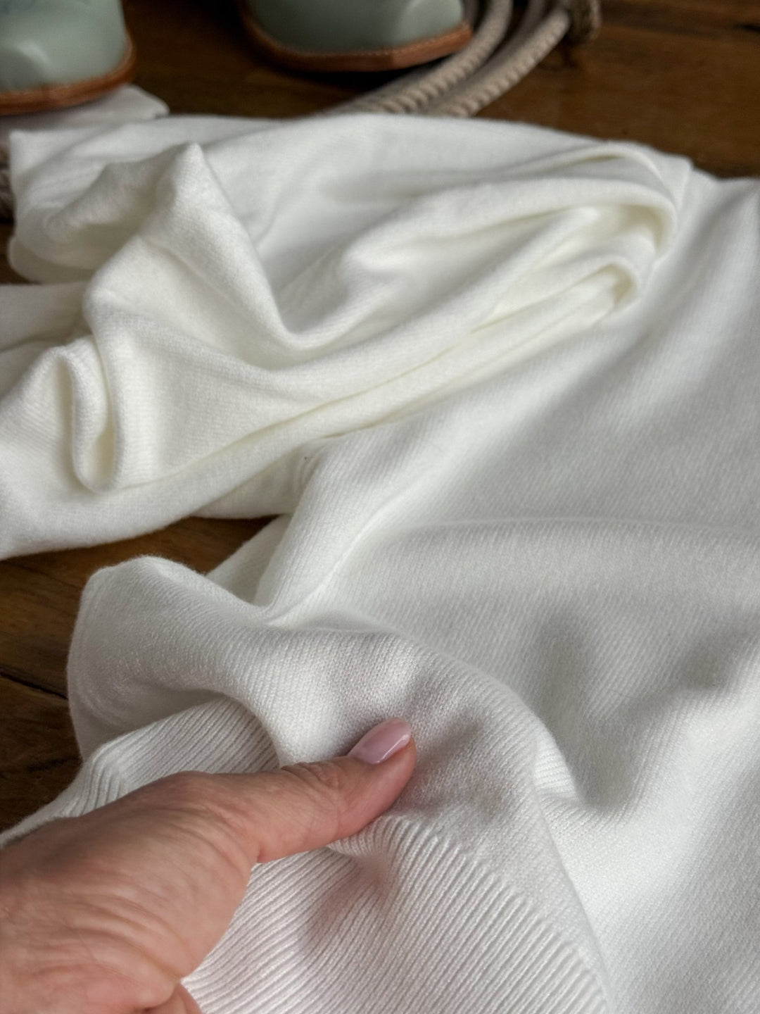 A hand with light pink nail polish touches the Arietta White Puff Sleeve Top by Savannah Sevens western life{&}style, which is spread out on a wooden surface. In the background, a pair of green shoes and a rope are partially visible.
