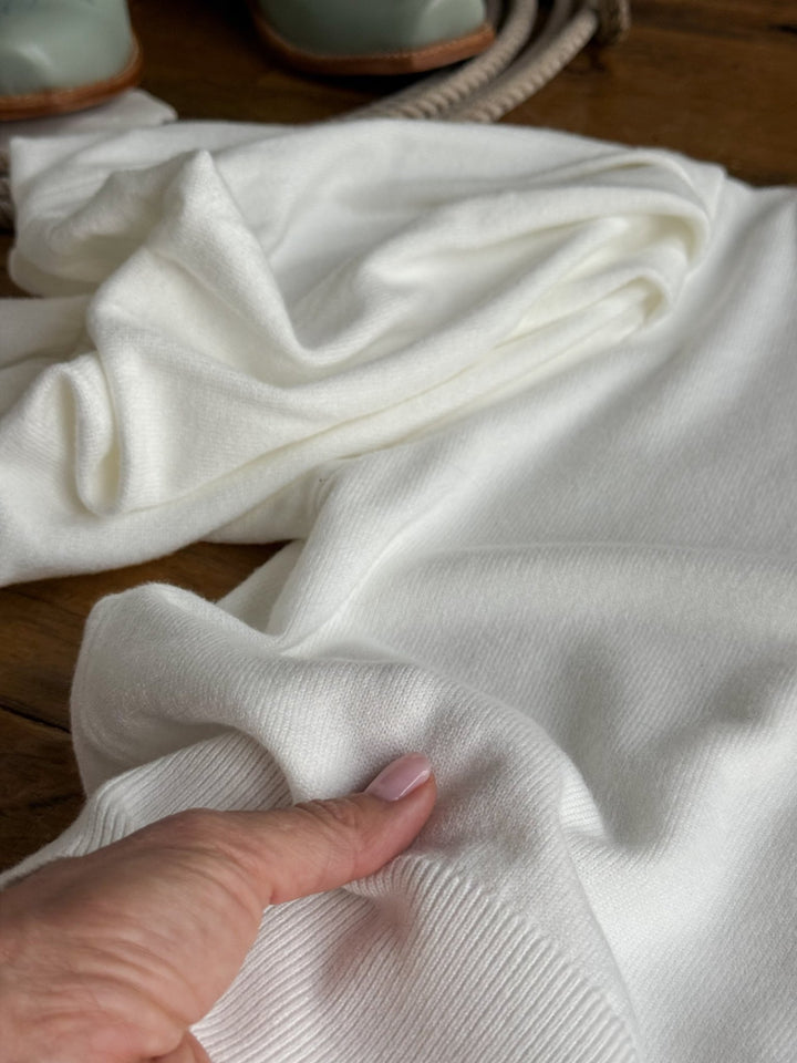 A hand with light pink nail polish touches the Arietta White Puff Sleeve Top by Savannah Sevens western life{&}style, which is spread out on a wooden surface. In the background, a pair of green shoes and a rope are partially visible.