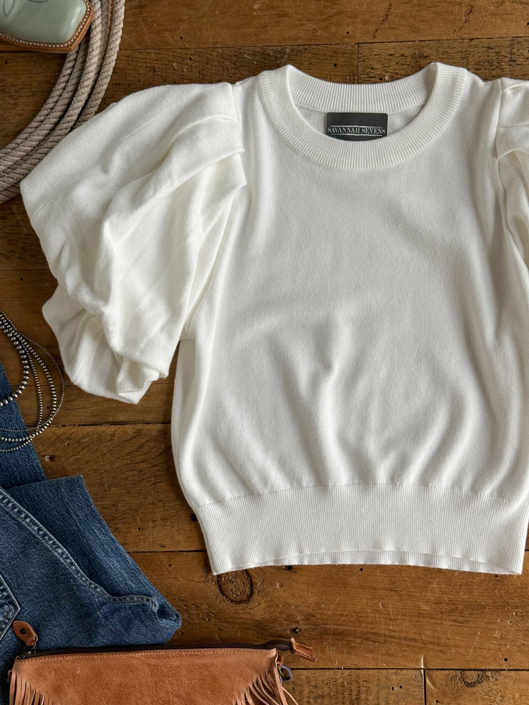 The Arietta White Puff Sleeve Top by Savannah Sevens western life{&}style is displayed flat on a wooden surface, accompanied by denim, a leather bag, and accessories.