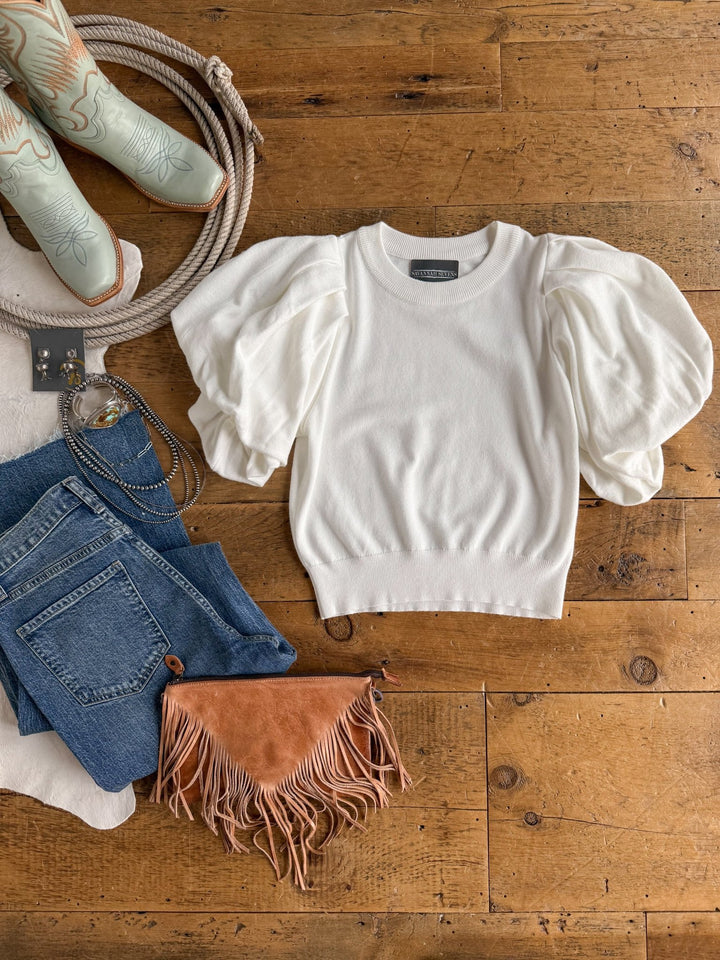 The Arietta White Puff Sleeve Top by Savannah Sevens western life{&}style is displayed on a wooden surface alongside blue jeans, a brown fringed clutch, a belt, turquoise cowboy boots, and silver jewelry.