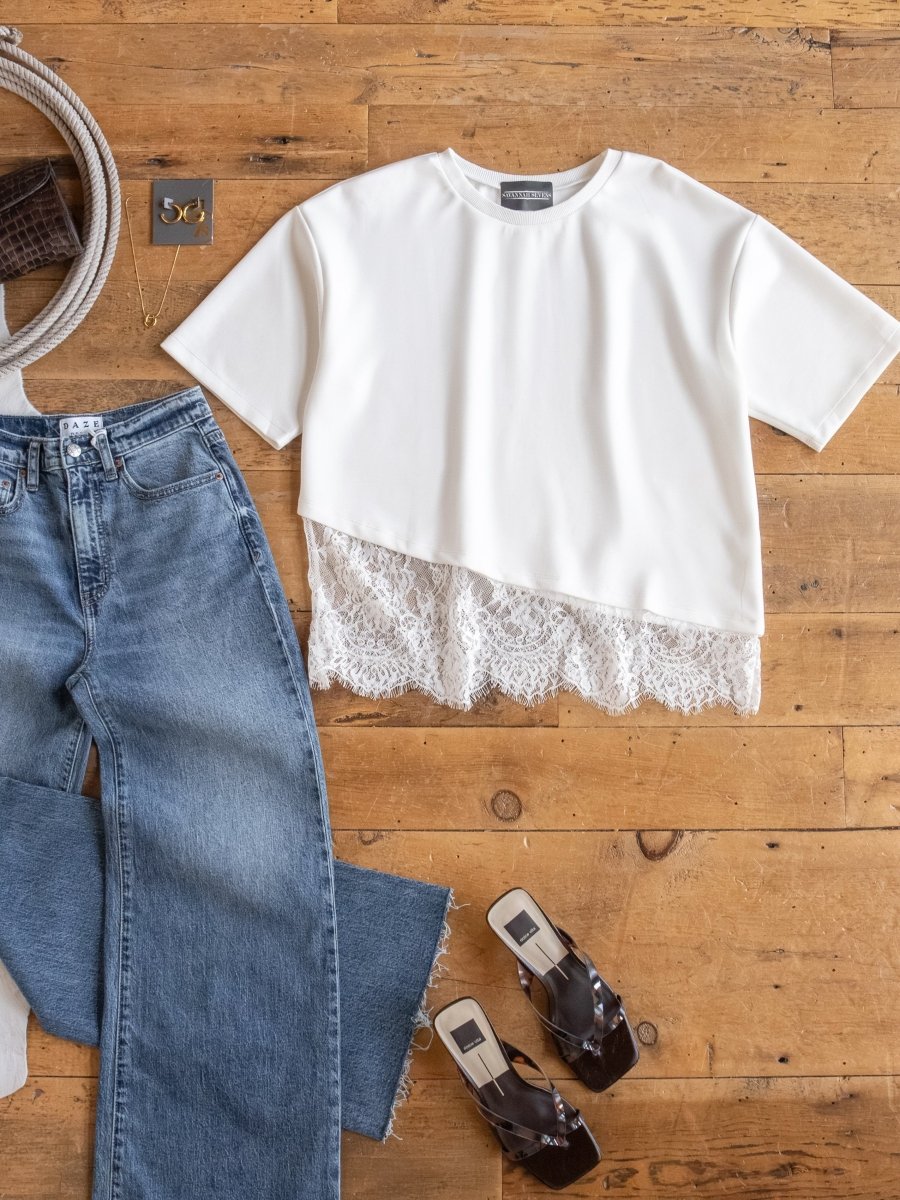 Flat lay of a casual outfit on a wooden floor featuring the Arivaca White Lace Hem Top by Savannah Sevens western life{&}style, blue wide-leg jeans, brown transparent-strap sandals, gray belt, gray fabric, and jewelry.