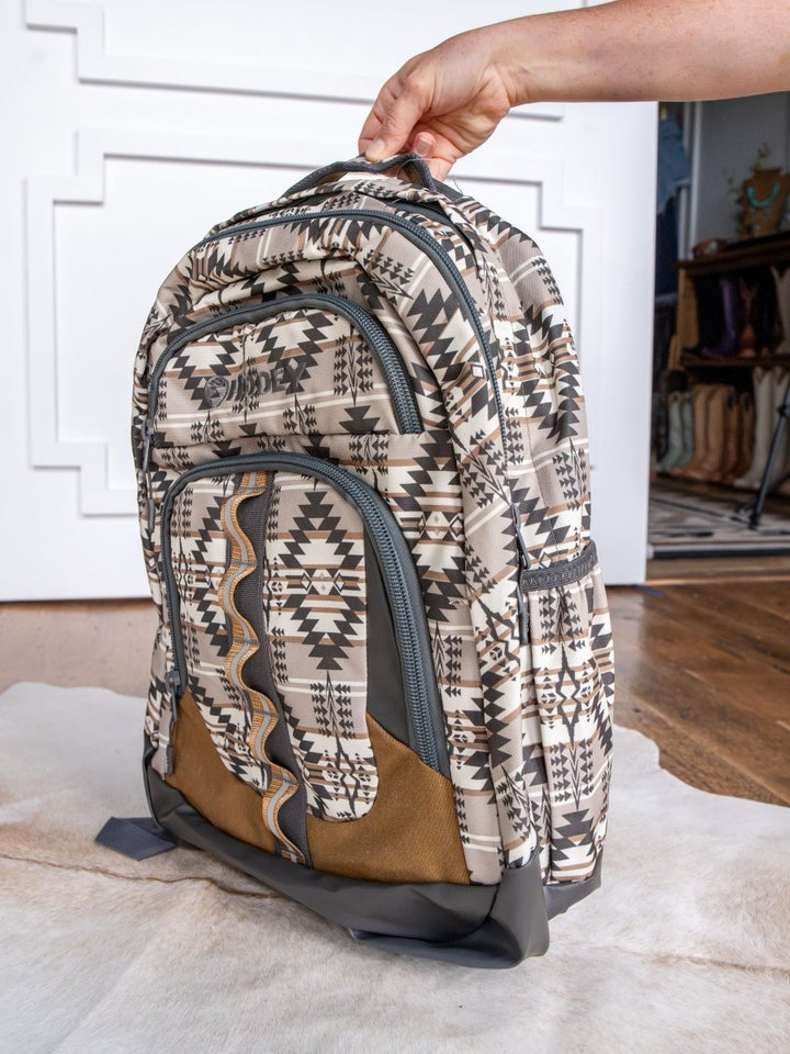 A hand holds up the Savannah Sevens western life{&}style Arizona Large Southwest Hat Holder Backpack, featuring geometric beige, black, and white designs with multiple zippered compartments. The background has a white wall and wooden floor.