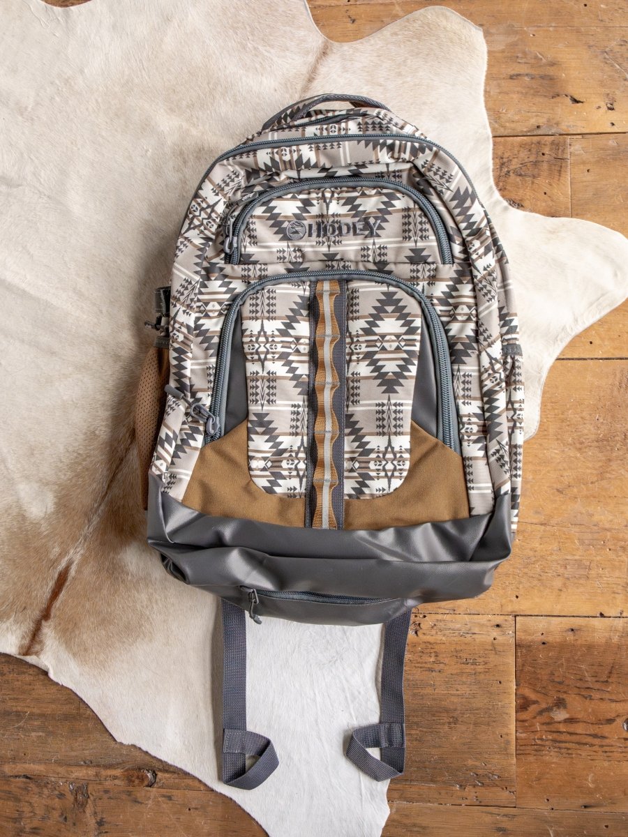 The Arizona Large Southwest Hat Holder Backpack by Savannah Sevens western life{&}style features geometric patterns in gray, beige, and brown with multiple zippered compartments and brown accents, shown on a white animal hide on a wooden floor.
