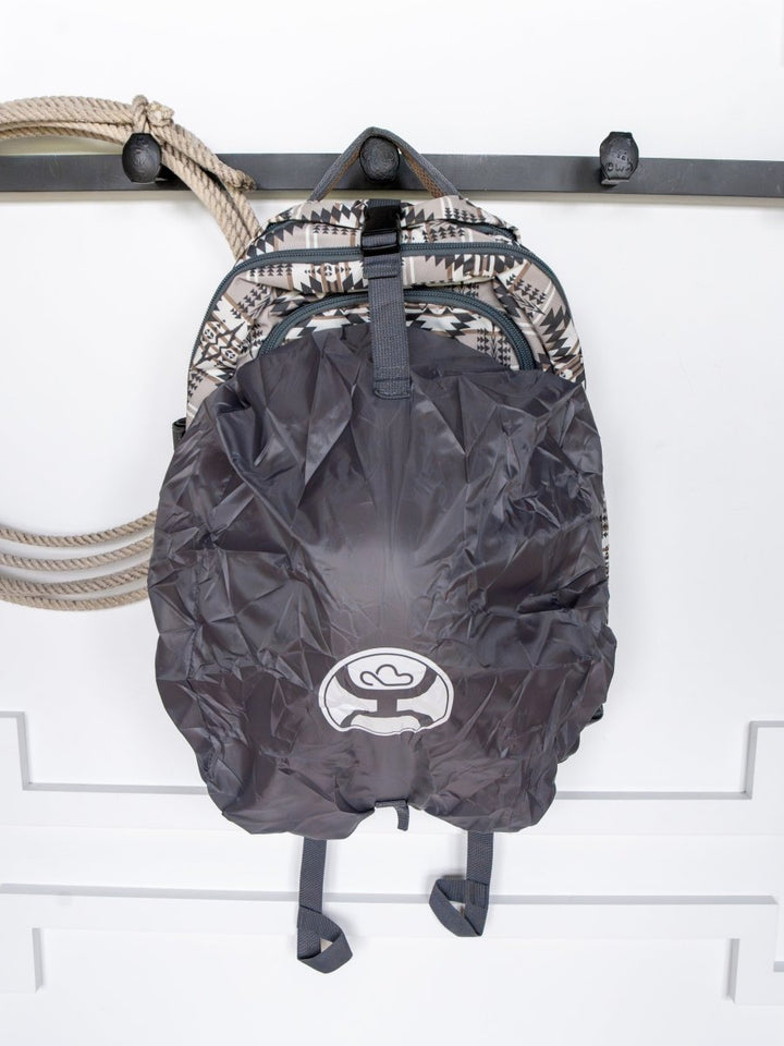 The Arizona Large Southwest Hat Holder Backpack by Savannah Sevens western life{&}style, with a patterned waterproof design and dark gray rain cover featuring a white logo, hangs on a wall hook next to a coiled rope.