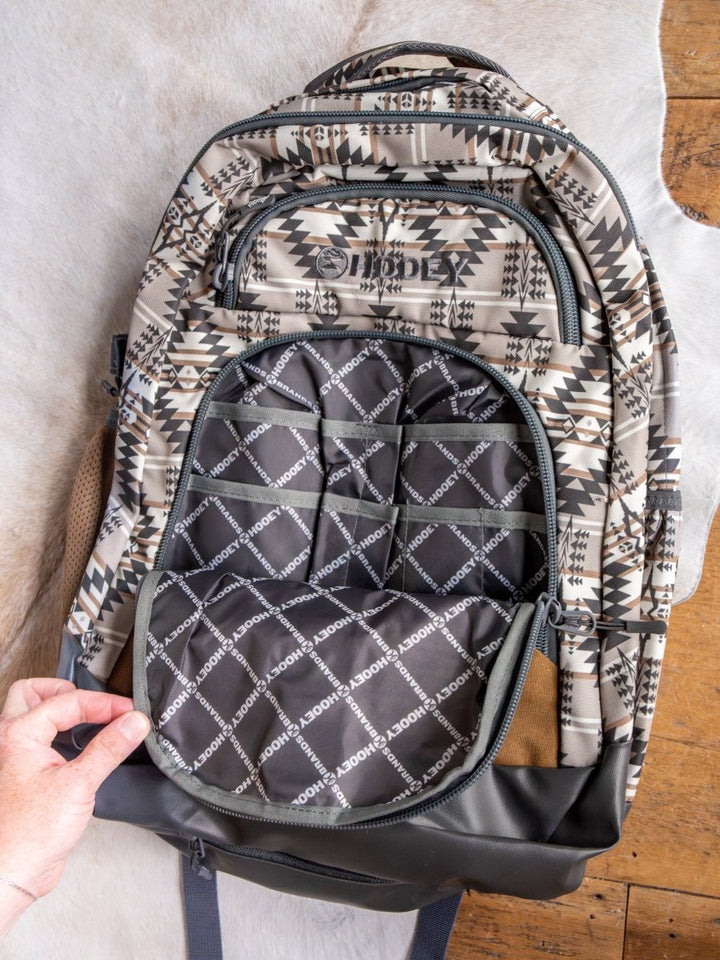 A hand opens the front pocket of the Arizona Large Southwest Hat Holder Backpack by Savannah Sevens western life{&}style, showing its black lining with a white geometric pattern and repeated branding. The backpack sits on a light surface above a wooden floor.