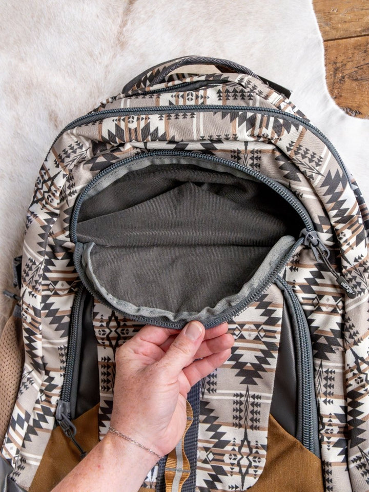 A hand opens the front zippered pocket of the Savannah Sevens western life{&}style Arizona Large Southwest Hat Holder Backpack, showcasing its empty interior and beige, black, and brown geometric pattern against a light background.