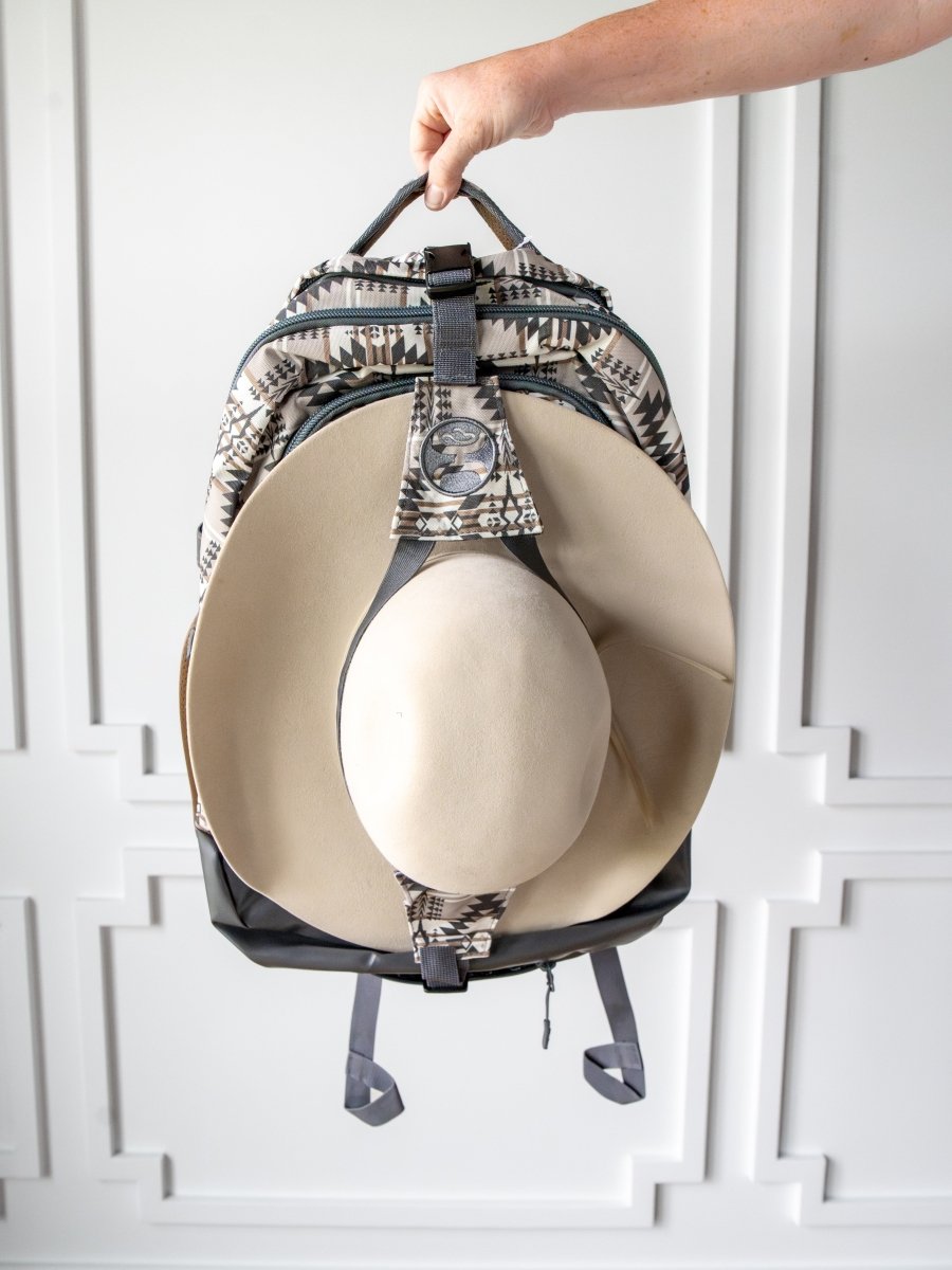 A hand holds the Savannah Sevens western life{&}style Arizona Large Southwest Hat Holder Backpack with a wide-brimmed beige hat attached, set against a white wall with decorative molding.