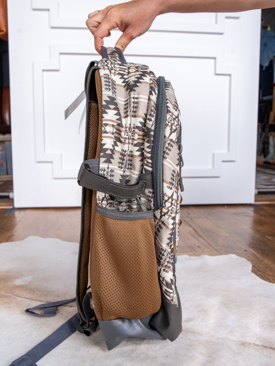 A hand holds the top handle of the Arizona Large Southwest Hat Holder Backpack by Savannah Sevens western life{&}style, standing upright on a cowhide rug with its side profile showing a brown mesh pocket and geometric design.