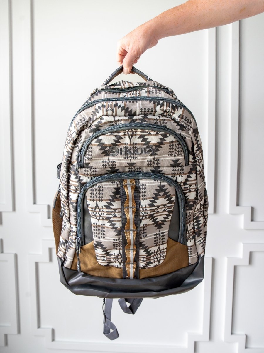 A hand holds the Arizona Large Southwest Hat Holder Backpack by Savannah Sevens western life{&}style, featuring geometric black, white, and brown designs with multiple zippered compartments, set against a white paneled background.