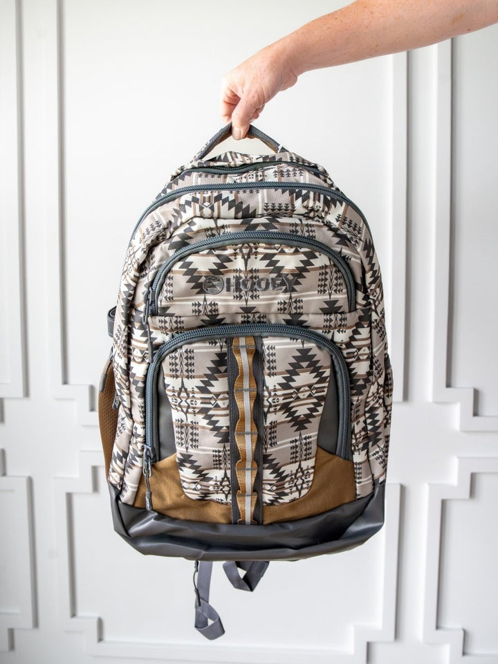 A hand holds the Arizona Large Southwest Hat Holder Backpack by Savannah Sevens western life{&}style, featuring geometric black, white, and brown designs with multiple zippered compartments, set against a white paneled background.