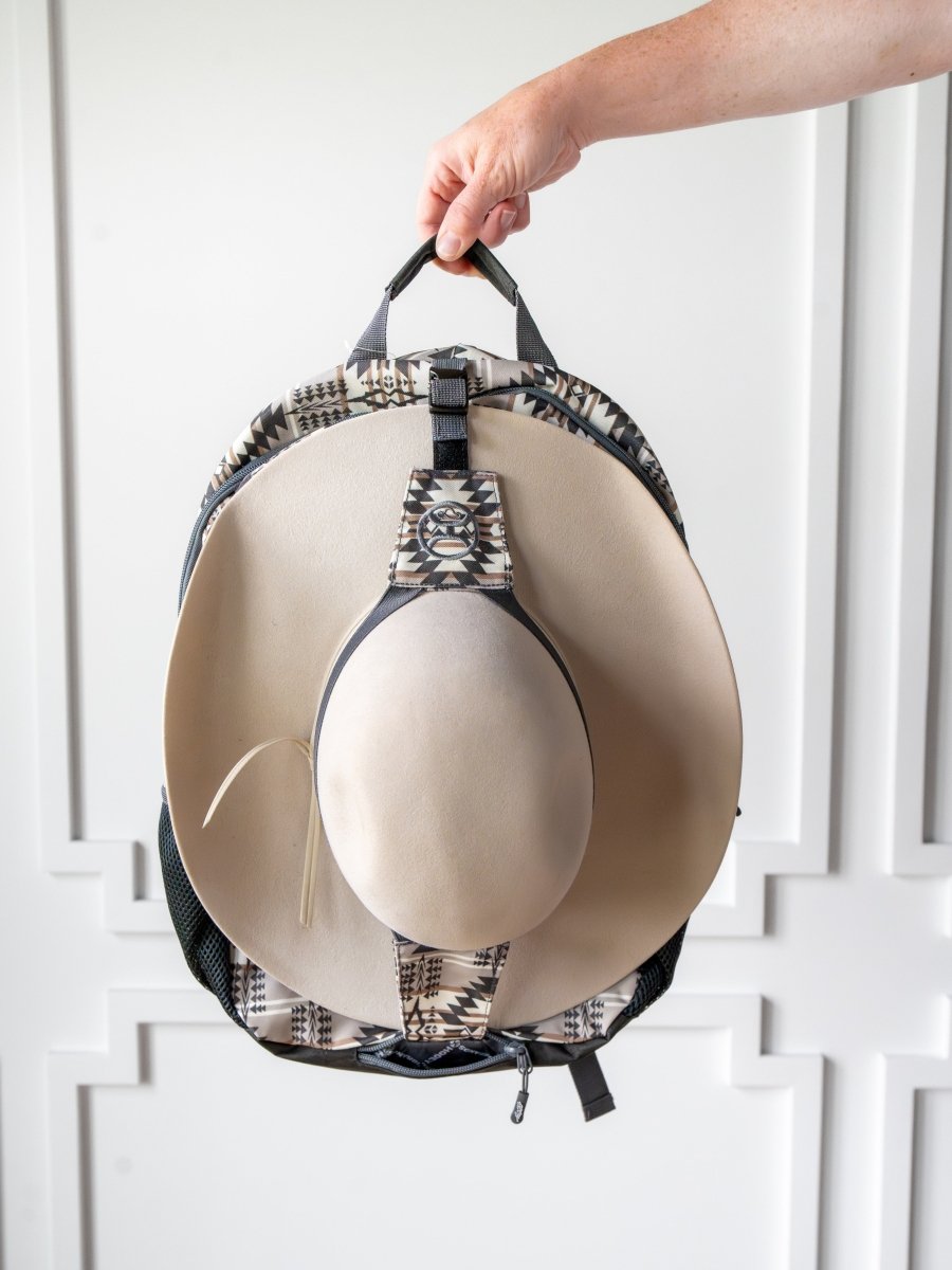 A hand holds the Arizona Medium Southwest Hat Holder Backpack by Savannah Sevens western life{&}style, with a Hooey hat holder securing a beige wide-brimmed hat to the front, set against a white background with decorative molding.