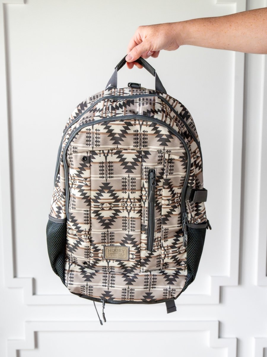 A hand displays the Savannah Sevens western life{&}style Arizona Medium Southwest Hat Holder Backpack, featuring black and beige geometric designs, two mesh side pockets, a laptop sleeve, and several zippered compartments.