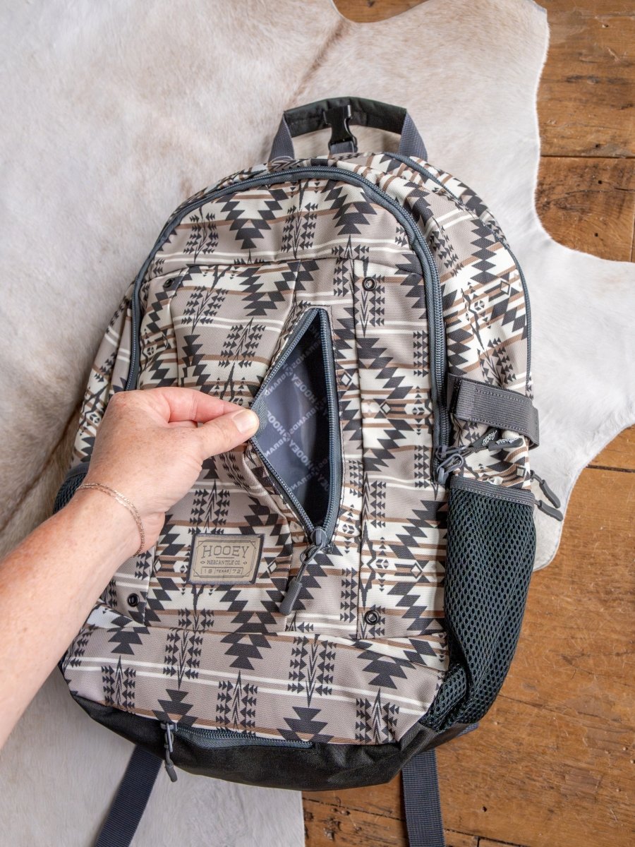 A hand opens a zippered pocket on the Savannah Sevens western life{&}style Arizona Medium Southwest Hat Holder Backpack, featuring geometric designs and a laptop sleeve, set on a light cowhide rug and wooden floor.