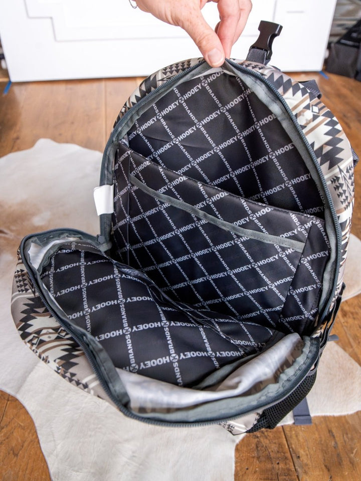 A hand opens the Savannah Sevens western life{&}style Arizona Medium Southwest Hat Holder Backpack, displaying its black interior with a white crisscross grid and "HOOEY" print, laptop sleeve visible, on a wooden floor above a white rug.