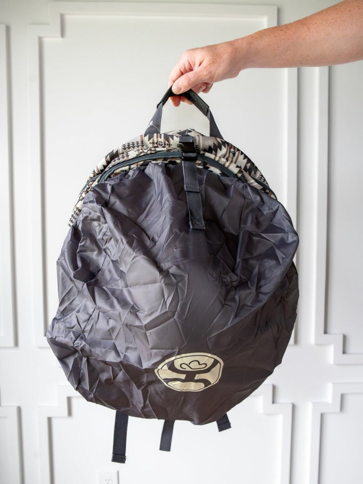 A hand displays the Savannah Sevens western life{&}style Arizona Medium Southwest Hat Holder Backpack, featuring a gray canvas with black straps and an abstract top pattern, in front of a white paneled wall.