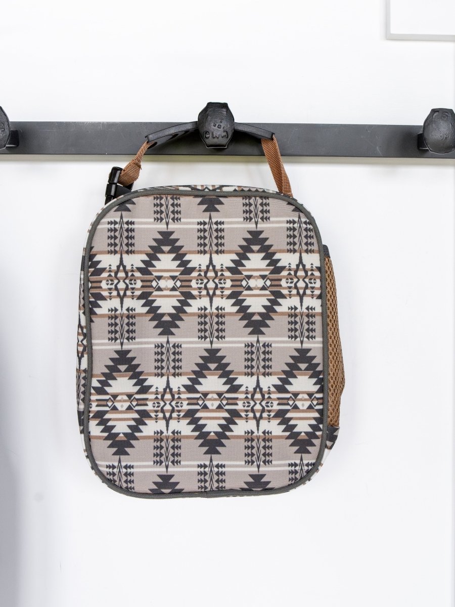 The Arizona Southwest Canvas Lunch Box by Savannah Sevens western life{&}style hangs from a black wall hook. With a geometric pattern, brown handle, mesh side pocket, and waterproof liner, it stands out against the white wall.