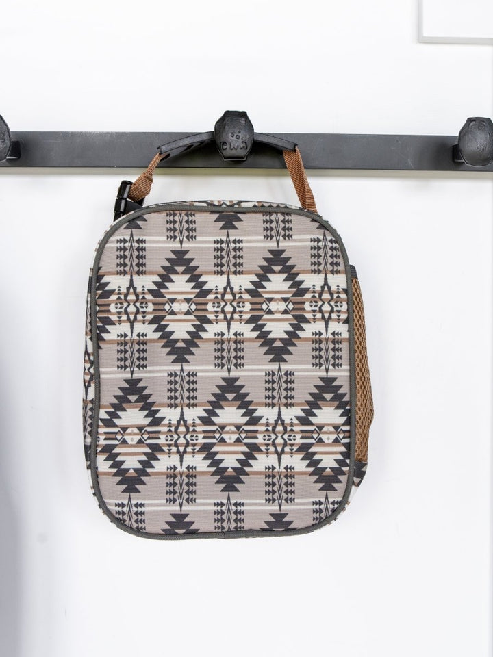 The Arizona Southwest Canvas Lunch Box by Savannah Sevens western life{&}style hangs from a black wall hook. With a geometric pattern, brown handle, mesh side pocket, and waterproof liner, it stands out against the white wall.