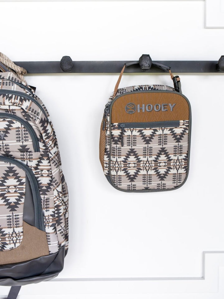 A patterned backpack and the Arizona Southwest Canvas Lunch Box from Savannah Sevens western life{&}style hang on a black wall hook. Both have geometric designs in gray, black, and brown against a white background.