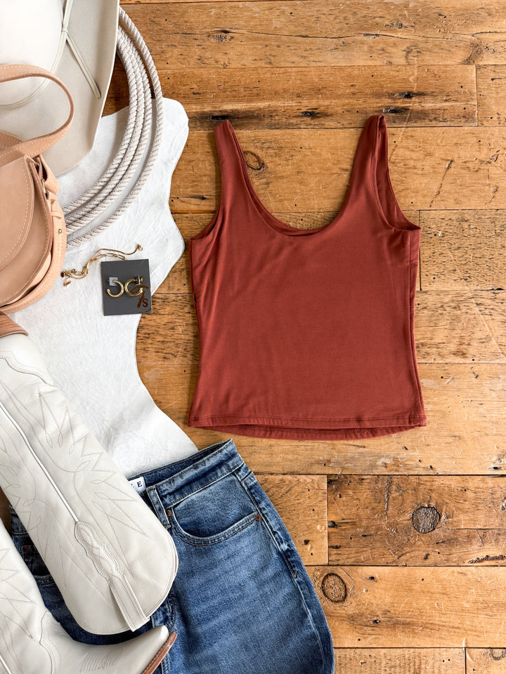 Flat lay of casual clothing and accessories on wood: the Arley Brick Scoop Neck Tank from Savannah Sevens western life{&}style, crafted from double-lined stretchy fabric, styled with blue jeans, white boots, a white top, a beige handbag, a coiled rope, and a pair of large gold hoop earrings.
