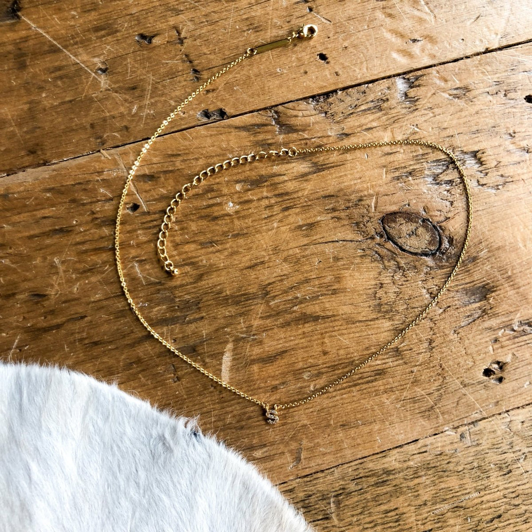 The Armada Initial Necklace by Savannah Sevens western life{&}style—a delicate gold piece with a small pendant—is displayed on a rustic wooden surface with white fur in the lower left corner.