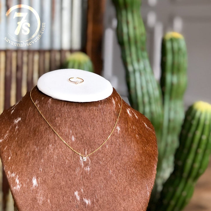 The Armada Initial Necklace by Savannah Sevens western life{&}style, featuring a gold “S” pendant, is displayed on a cowhide stand with a matching ring. A blurred green cactus and the Savannah Sevens logo are visible in the background.