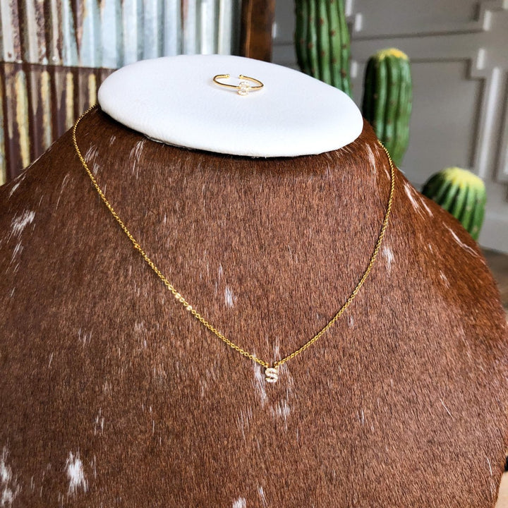 The Armada Initial Necklace by Savannah Sevens western life{&}style, featuring a gold “S” pendant, is displayed on a brown and white cowhide bust. A gold ring sits above on a white platform with green cacti and a rustic wall in the background.