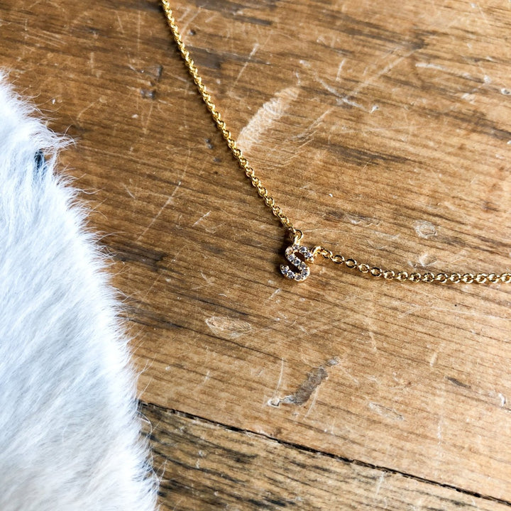 The Armada Initial Necklace by Savannah Sevens western life{&}style features a petite gold chain with a diamond-studded S pendant, displayed on a wooden surface beside a patch of white fur.