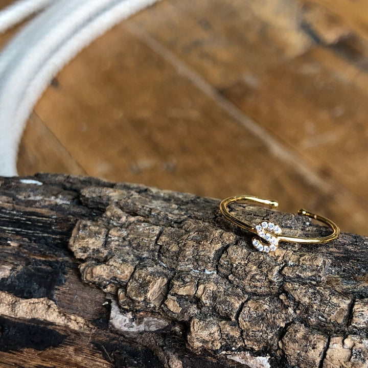 The Armada Initial Ring by Savannah Sevens western life{&}style features a gold band with clear stones forming the letter S, displayed on textured bark with wood and white rope in the background. 