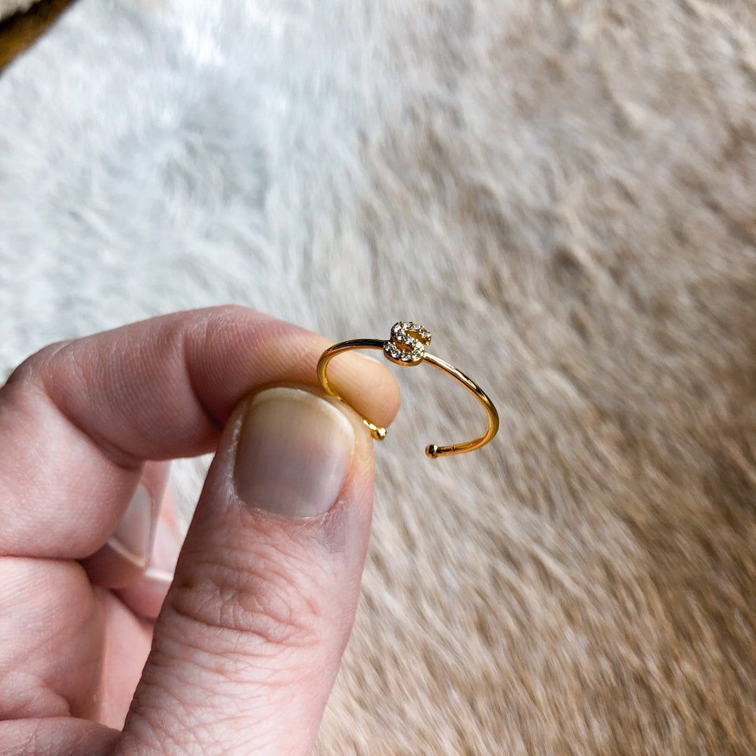 A hand displays the Armada Initial Ring by Savannah Sevens western life{&}style—a gold adjustable piece with a sparkling stone cluster—against a soft, light brown textured background.