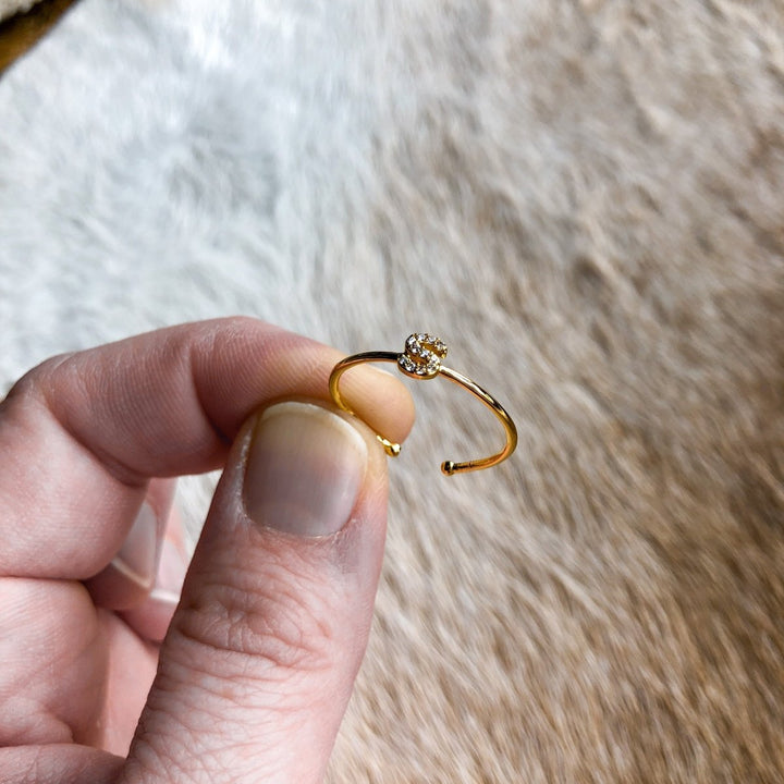 A hand displays the Armada Initial Ring by Savannah Sevens western life{&}style—a gold adjustable piece with a sparkling stone cluster—against a soft, light brown textured background.