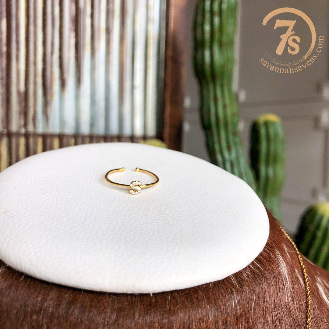 The Armada Initial Ring by Savannah Sevens western life{&}style, a delicate gold ring with a small clear stone, rests on a white cushion. A cactus and the savannahsevens.com logo appear in the background, showcasing this handmade jewelry piece.
