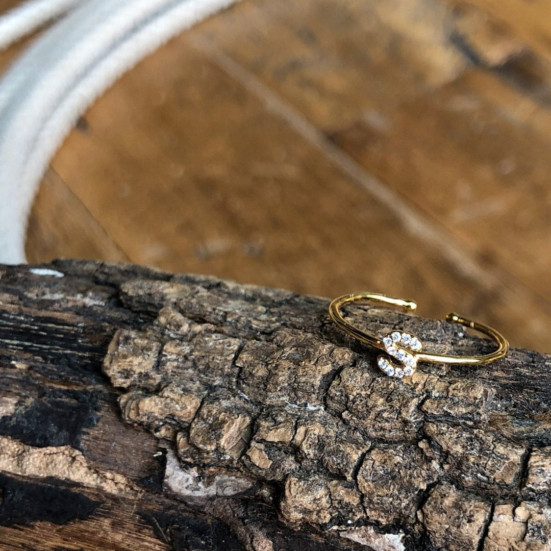The Armada Initial Ring by Savannah Sevens western life{&}style features a gold band with clear stones forming the letter S, displayed on textured bark with wood and white rope in the background. 