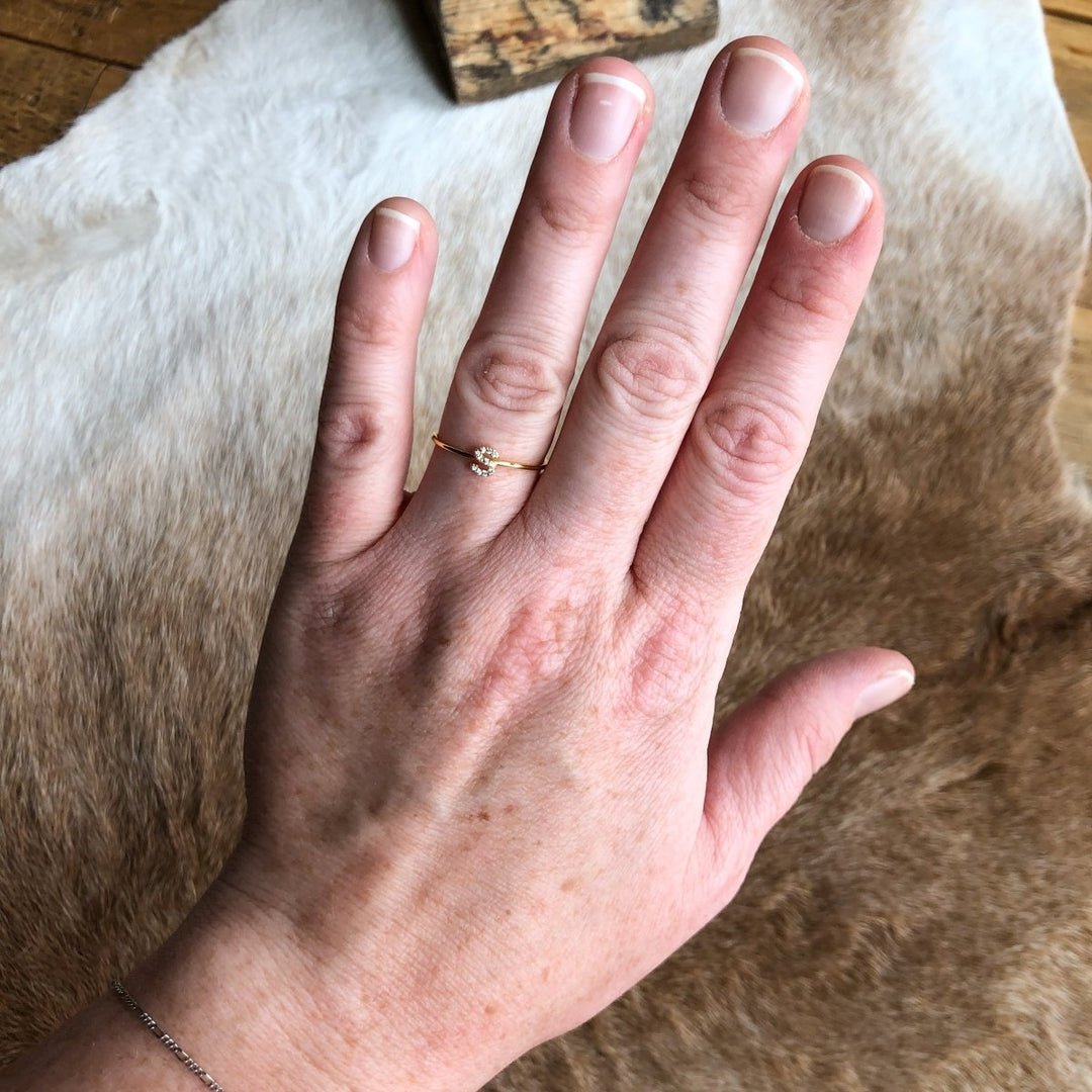 A hand with natural, unpolished nails displays the handmade Armada Initial Ring by Savannah Sevens western life{&}style, featuring a delicate gold design and small stone, resting atop a brown and white animal hide surface.