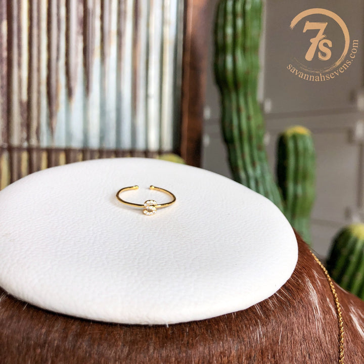 The Armada Initial Ring by Savannah Sevens western life{&}style, a delicate gold ring with a small clear stone, rests on a white cushion. A cactus and the savannahsevens.com logo appear in the background, showcasing this handmade jewelry piece.