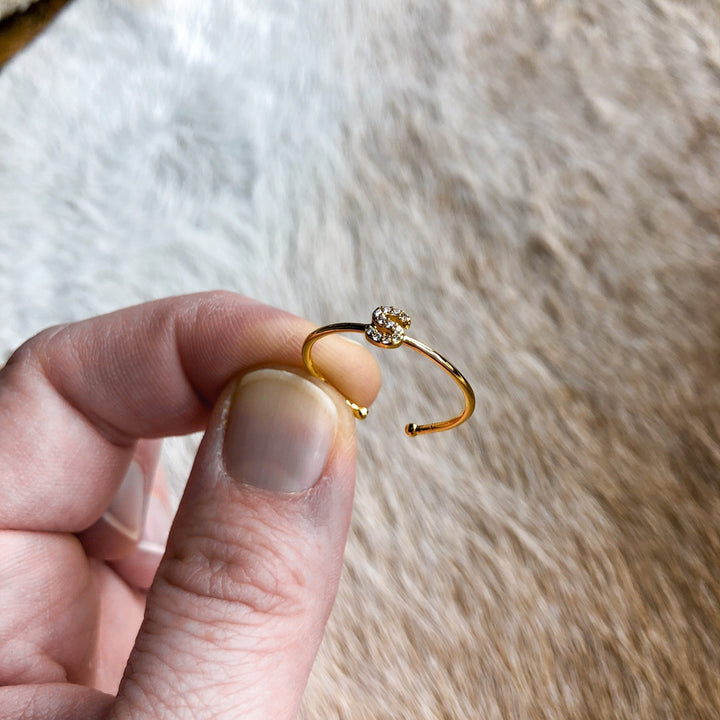 A hand displays the Armada Initial Ring by Savannah Sevens western life{&}style—a gold adjustable piece with a sparkling stone cluster—against a soft, light brown textured background.