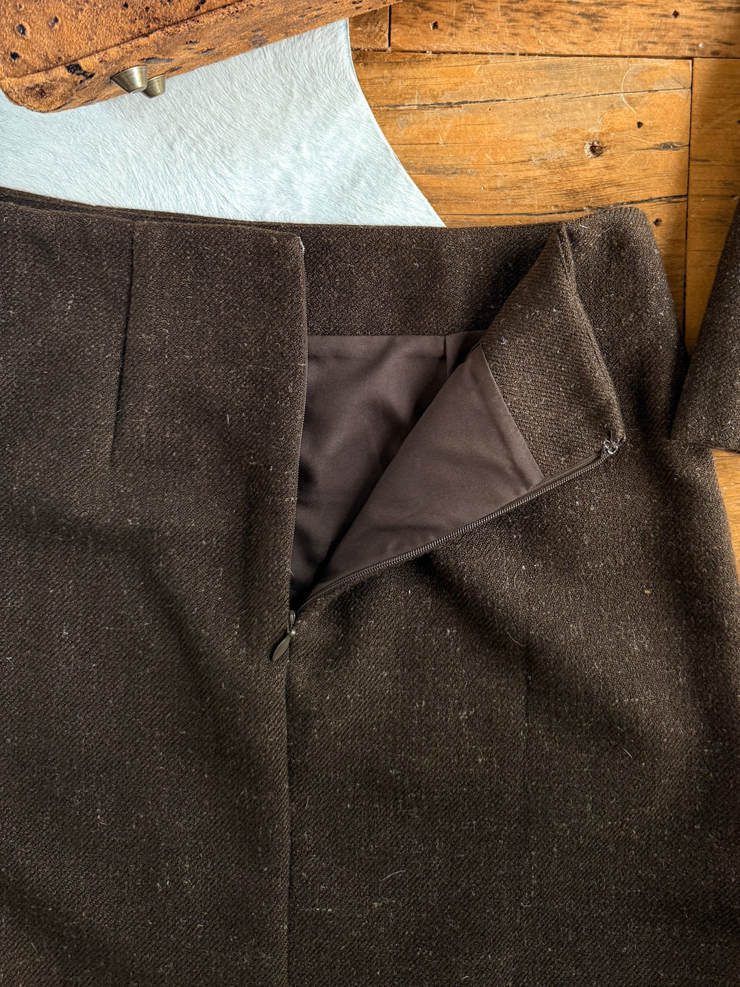 The Armona Chocolate Twill Vest Set from Savannah Sevens western life{&}style rests on a wooden surface, its dark brown wool pants partially unzipped to reveal the inner lining and zipper, displaying a high-waisted skirt-inspired design.